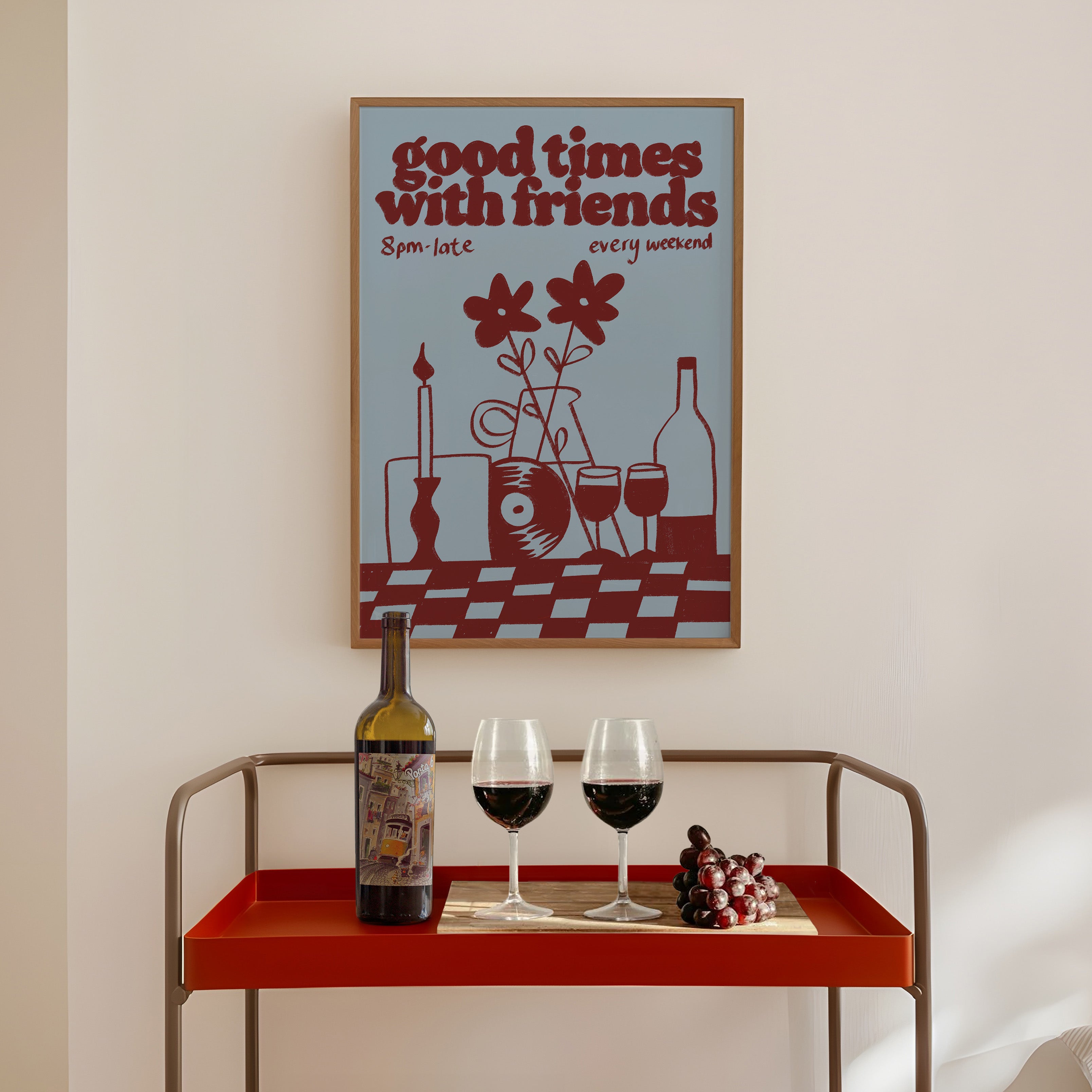 A framed retro-style wall art print with the text 'good times with friends' hangs above a red bar cart holding wine glasses, a wine bottle, and grapes in a softly lit interior.